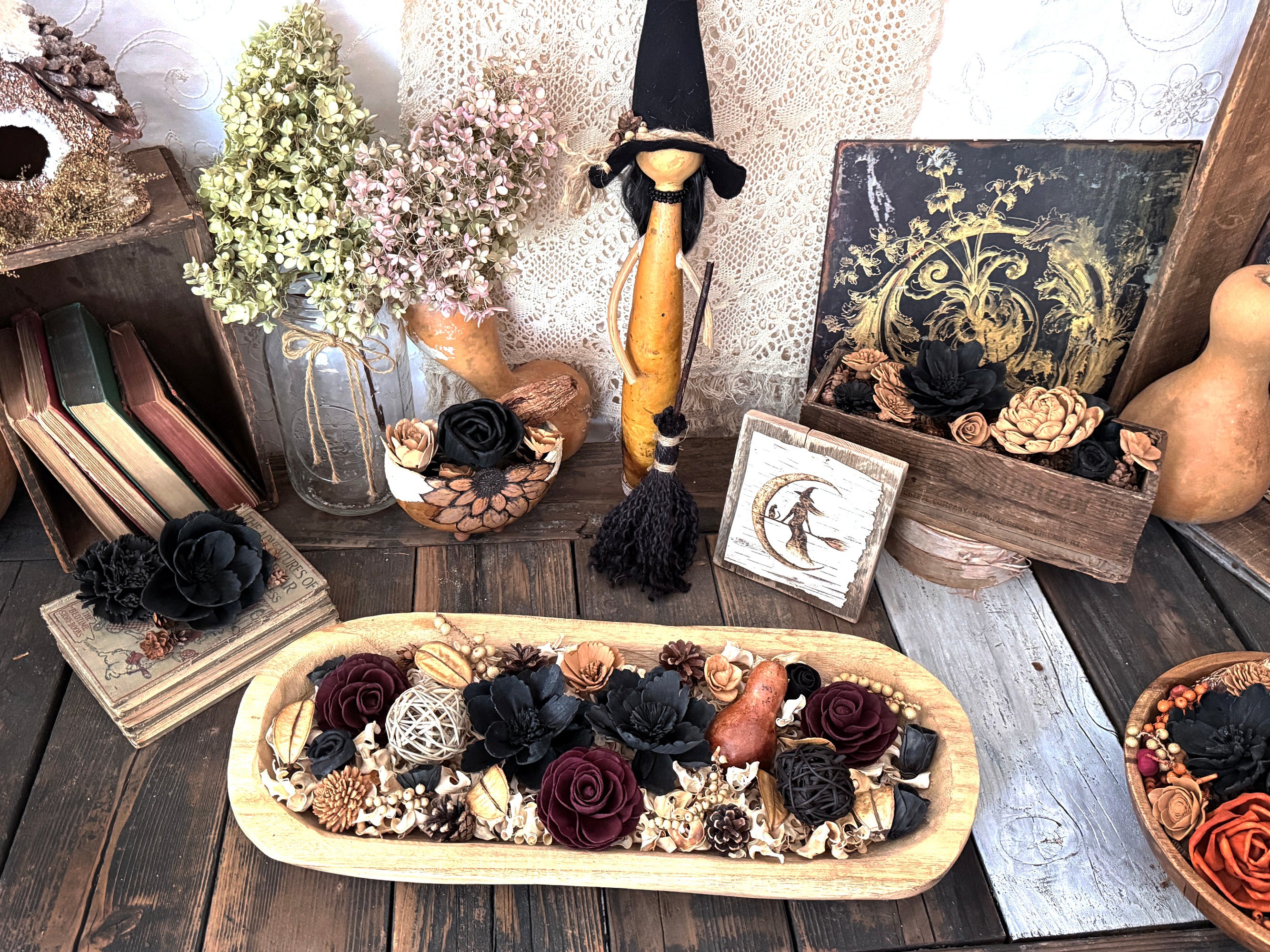 Decorative setup with a bowl of flowers, books, and a witch with her broom on a wooden surface.