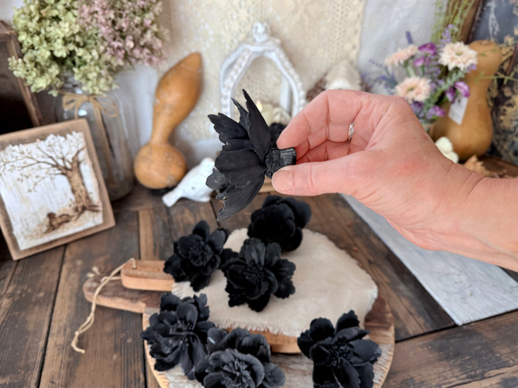 Hand holding a black flower blossom with profile view.  In the back are black flowers on a wooden surface with a floral and decorative background.