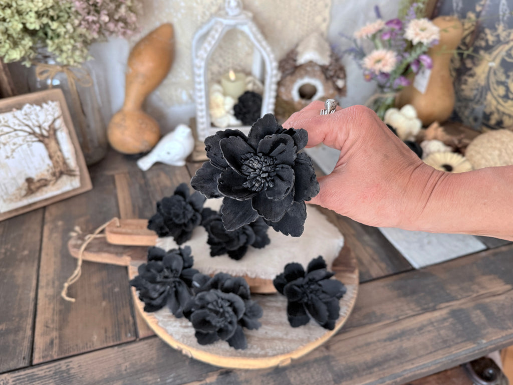 Hand holding a black flower for front view. An arrangement of more flower blossoms are on a wooden surface with decorative items in the background.