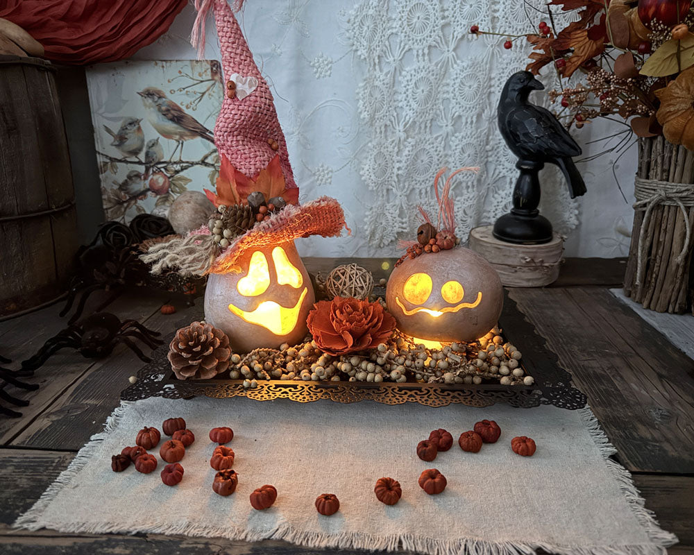 A Halloween table display showing a black tray with two lit gourds and decorative wood flowers a dried botanicals arranged around them. A black crow stands in the back.