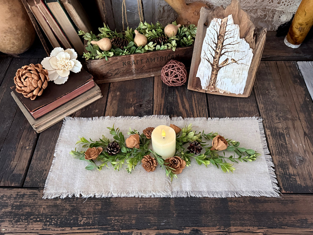 Decorative table runner setting with a candle, greenery, wood flower blossoms and pinecones on a linen table runner.