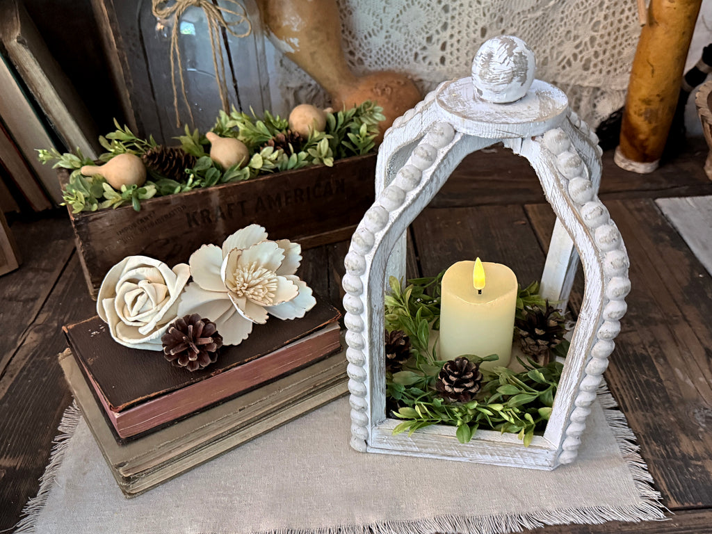 White beaded wood lantern holding candle and greenery. Vintage books stacked on the left with beige wood flowers sitting on top, 