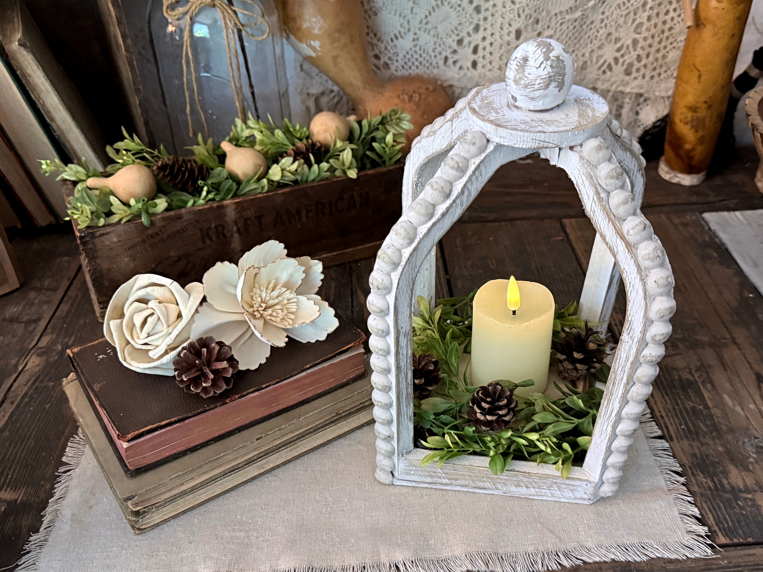 White beaded wood lantern holding candle and greenery. Vintage books stacked on the left with beige wood flowers sitting on top, 