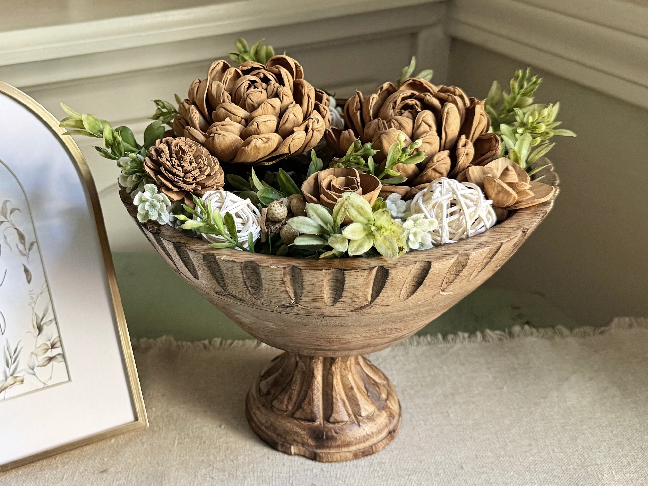 Teak wood bowl with riser holding boxwood greenery arrangement and wood flowers. 