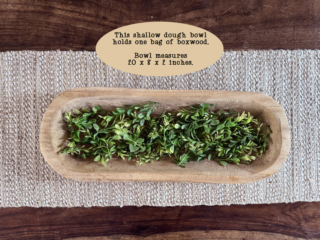 Wooden dough bowl filled with boxwood on a textured table runner on top of a coffee table. 