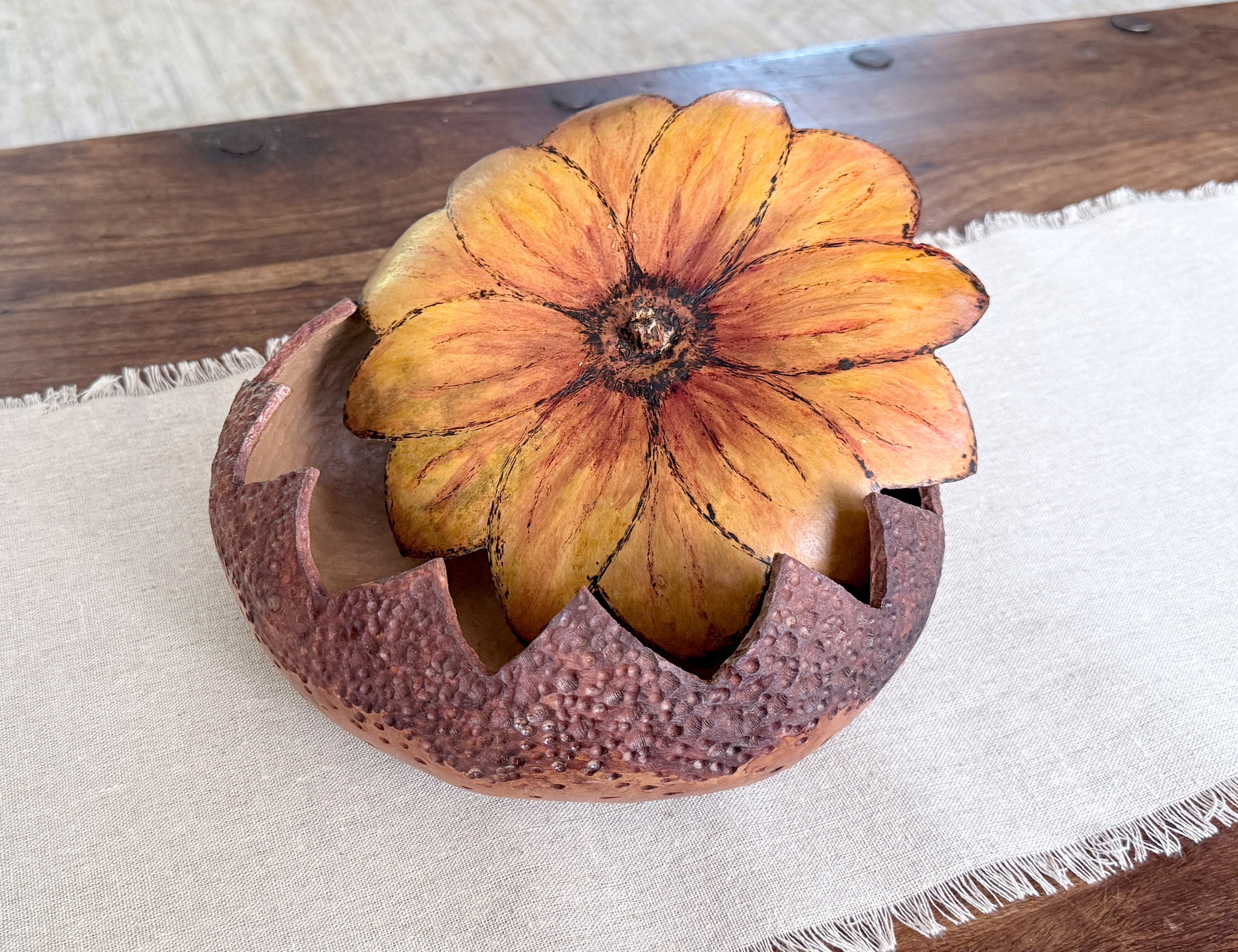 Hand carved gourd bowl with the sunflower blossom lid sitting askew. Bowl sits on linen runner on wood coffee table.