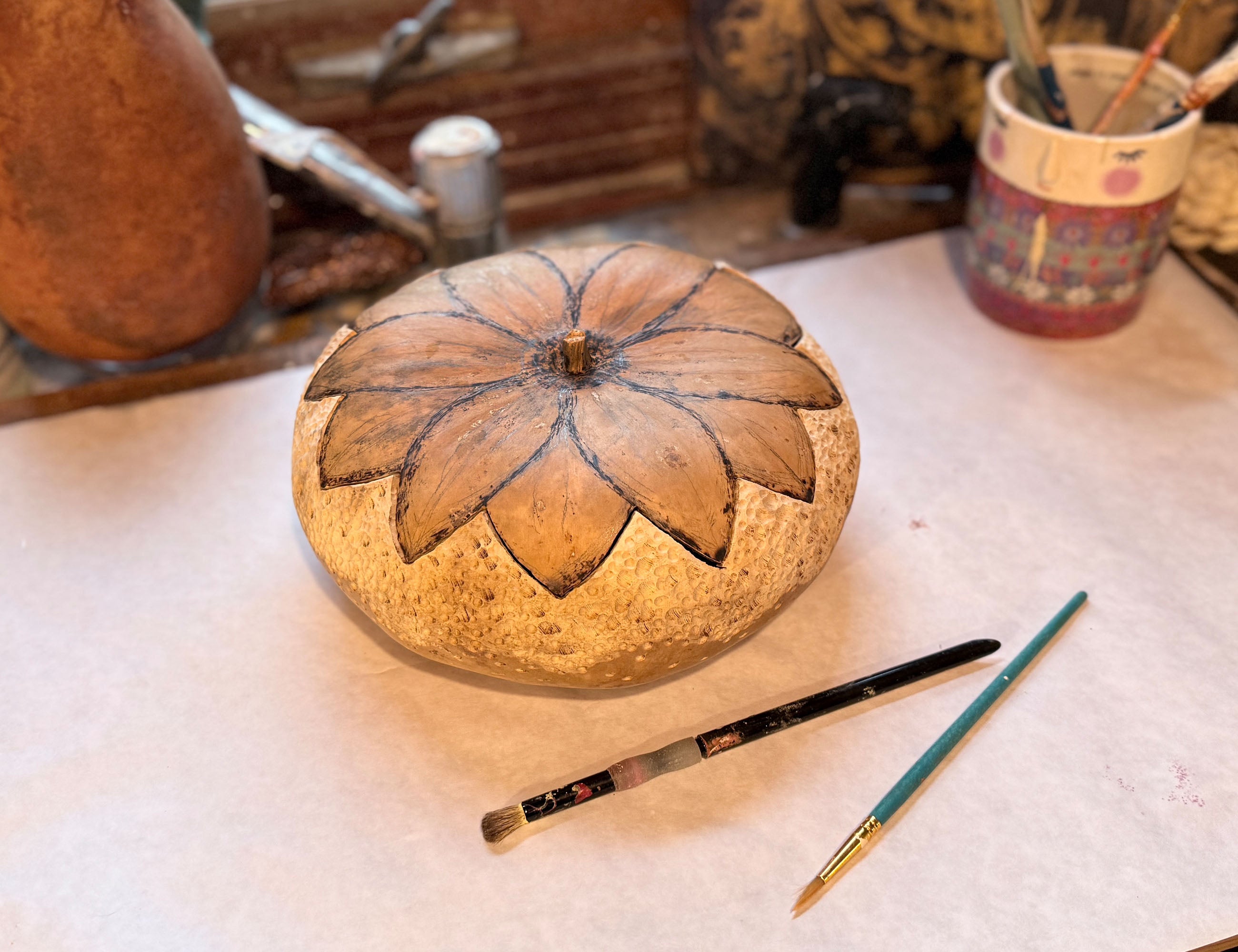 A carved gourd bowl sits on work station prior to being painted. Paint brushed are laid in front of gourd.