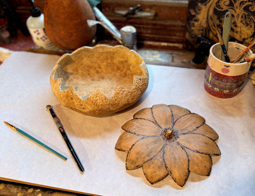 A round gourd sits on a work station with the carved flower lid sitting next to it. Paint brushes and supplies sit near by.