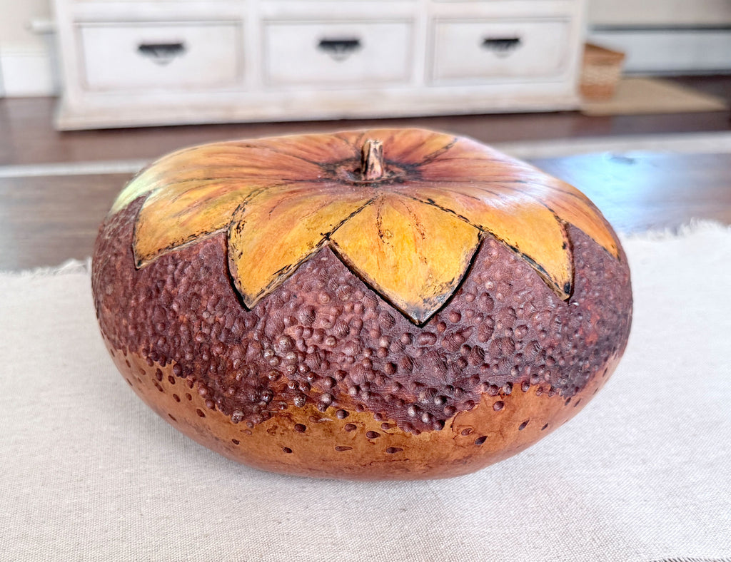 A decorative carved pumpkin with a textured dark brown base and large yellow-orange flower-like segments around the top, displayed on a light fabric surface with a blurred indoor background.