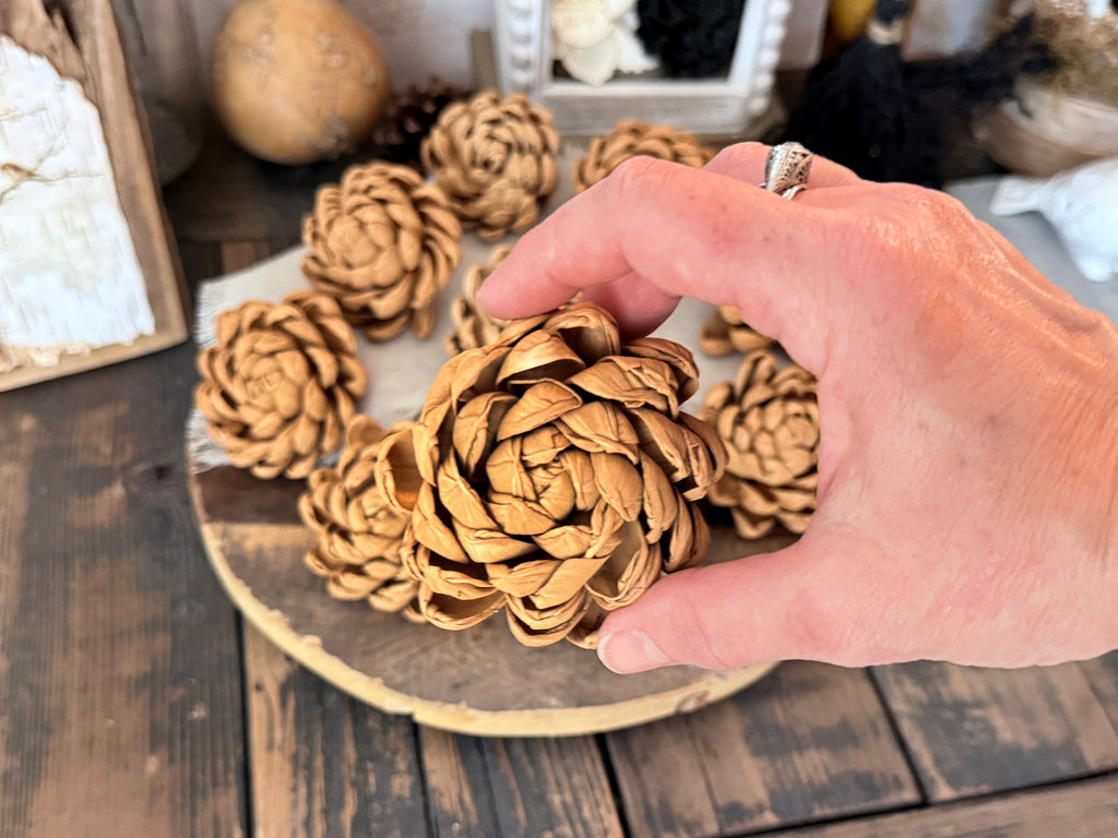 Hand holding a decorative wood flower on a wooden surface with more pine cones in the background.