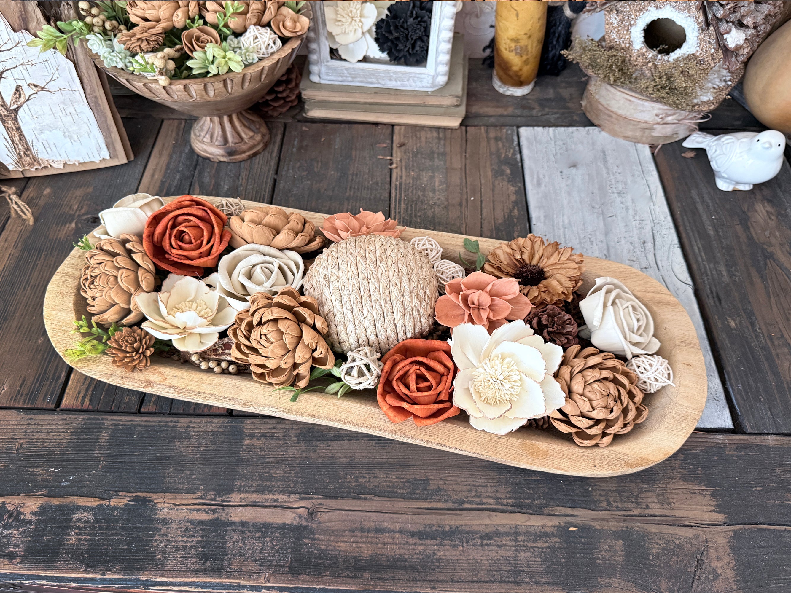 Decorative dowl bowl arrangement of orange, brown, and cream colored flowers and pinecones on a wooden tray with a rustic background.