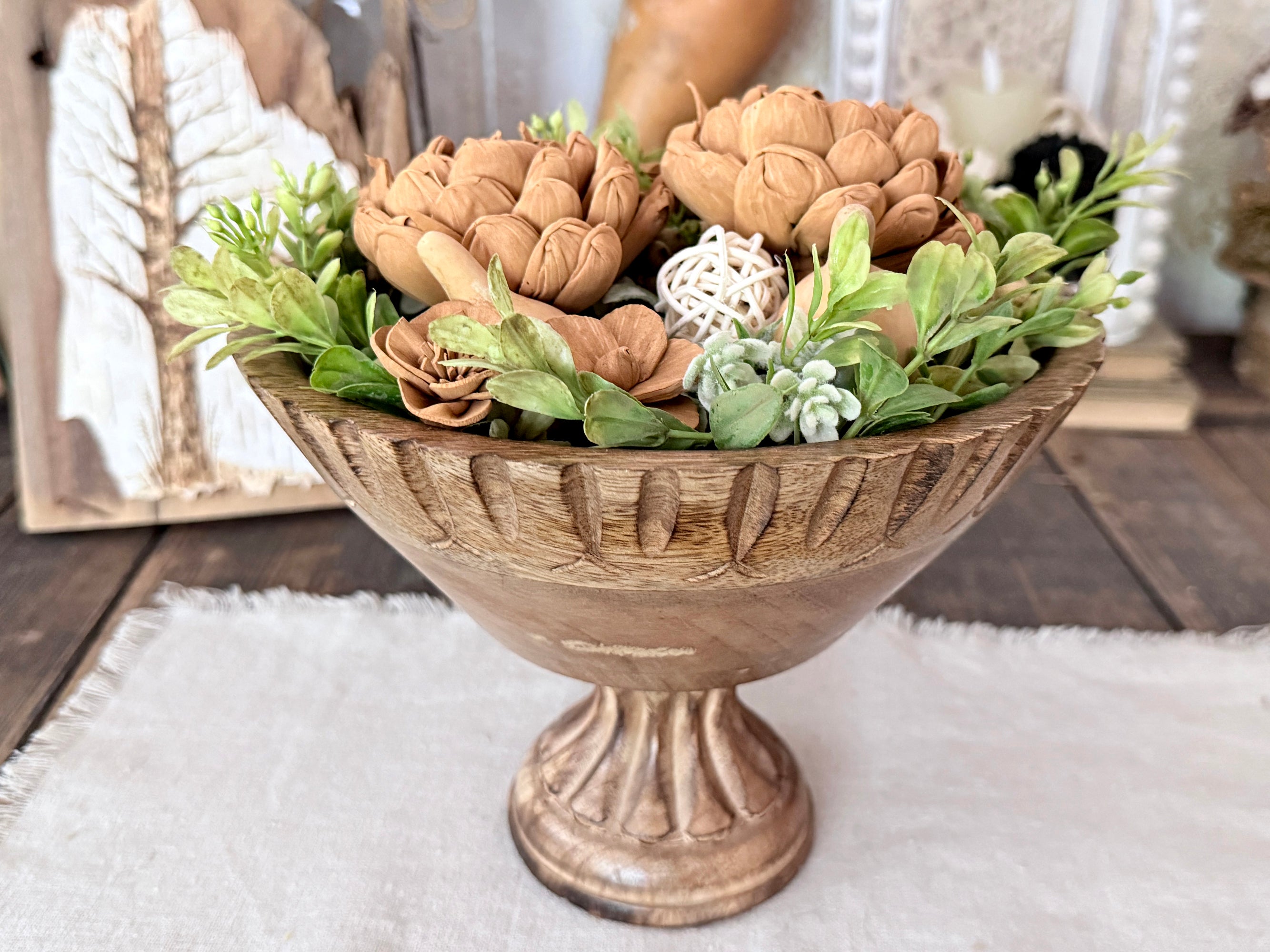 Decorative teak wood bowl with a riser holding  artificial wood flowers and plants on a wooden table.