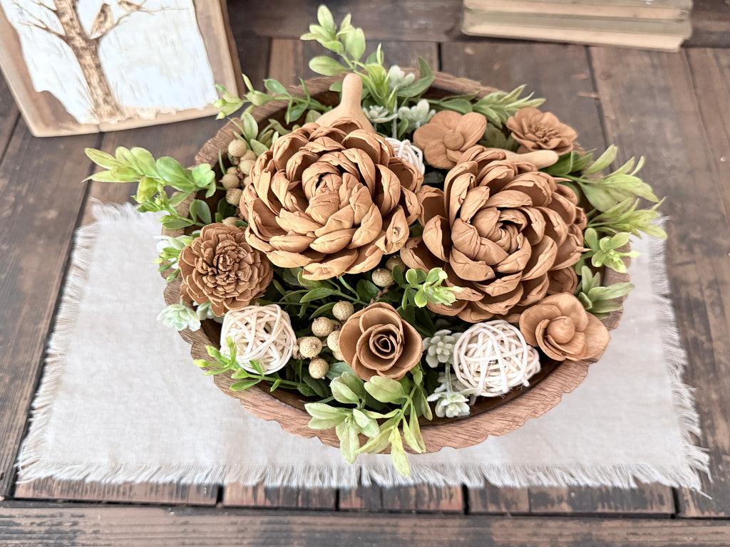 Decorative arrangement of wood flowers and greenery in a wooden bowl on a wooden surface.