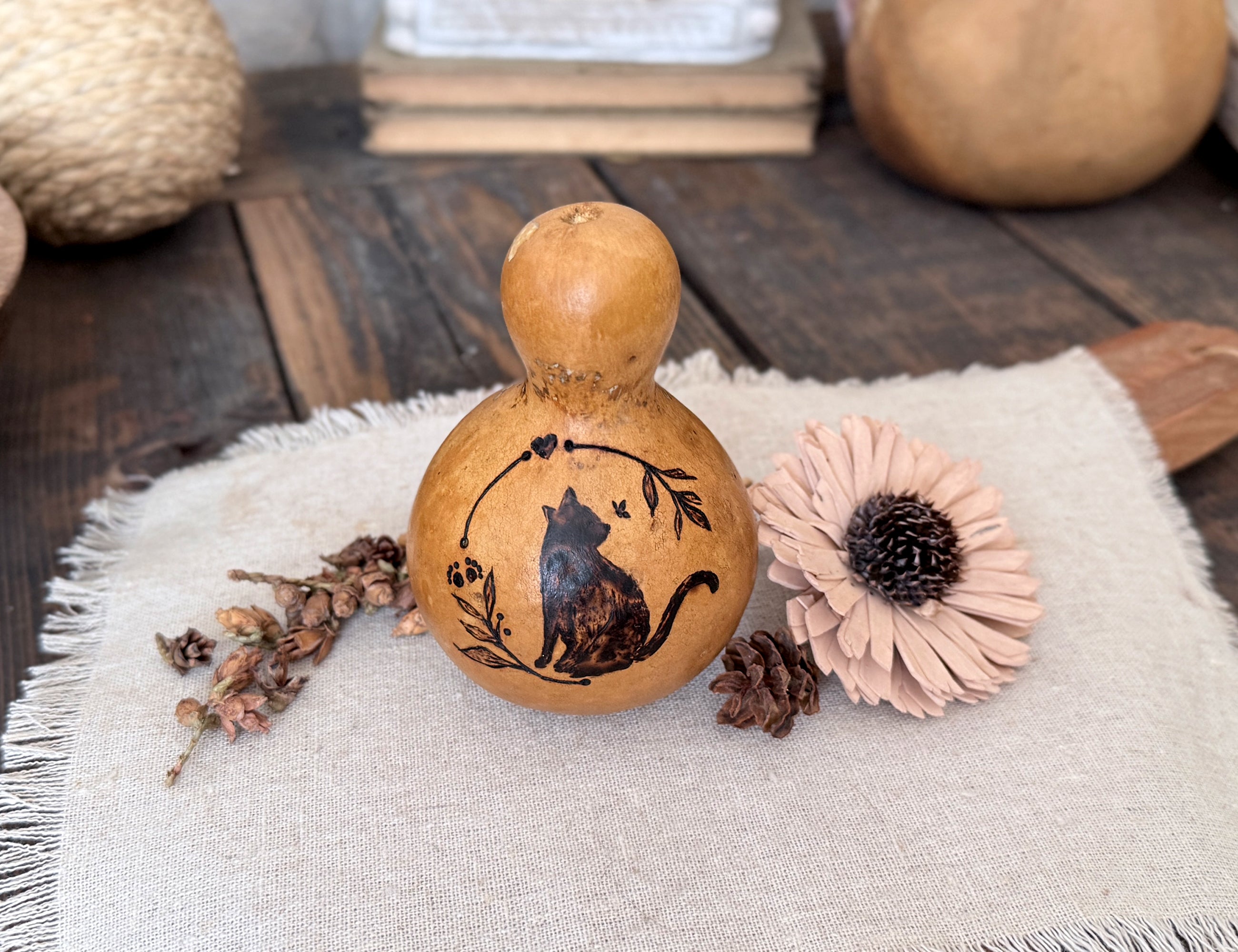 Decorative gourd with a cat design on a textured surface with flowers and pinecones.