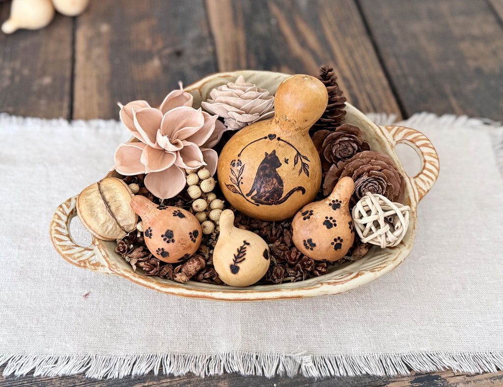 Decorative cat gourd is nestled in with smaller pet themed gourds, woods flowers, dried botanicals and twig ball in a ceramic bowl with handles. 