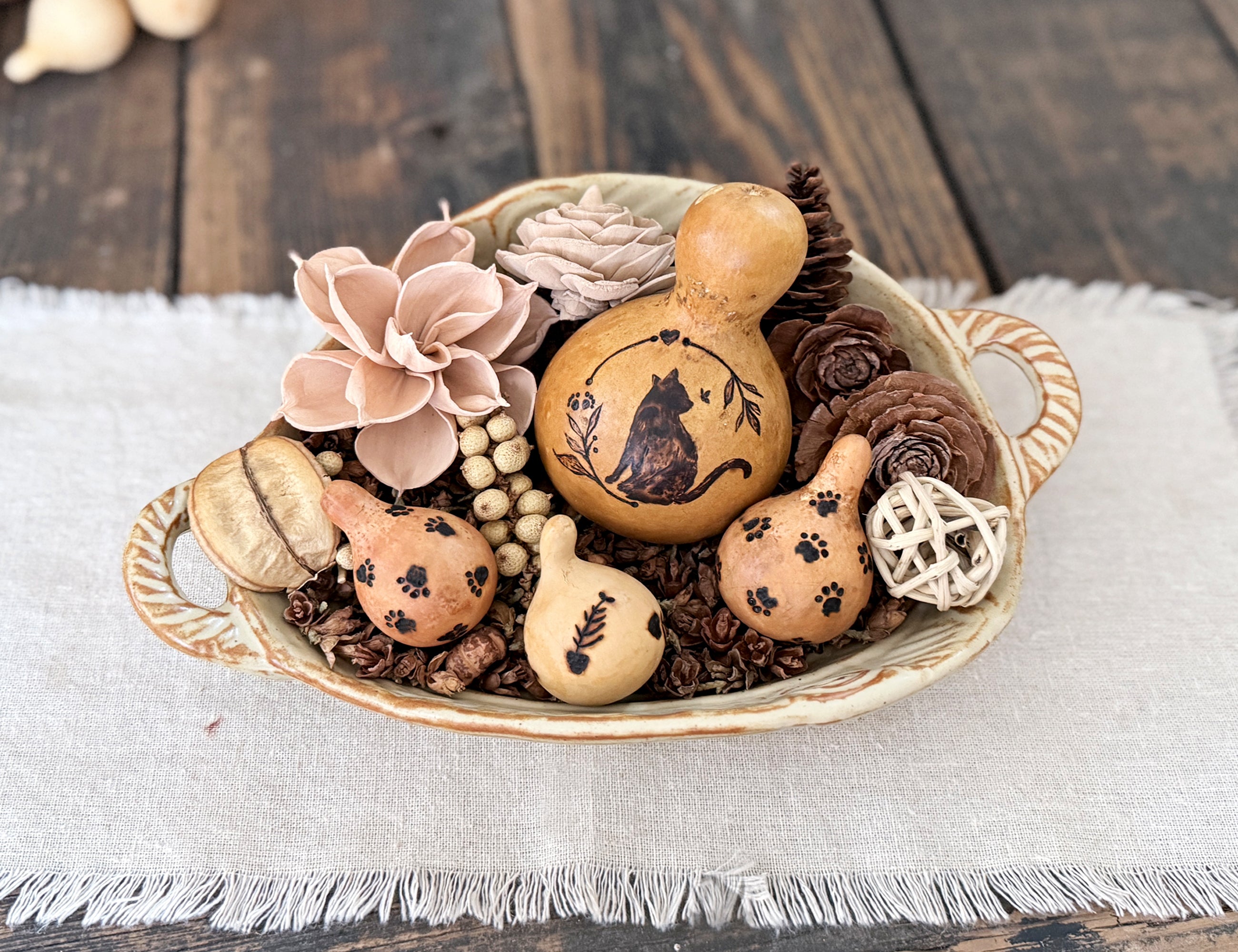 Decorative cat gourd is nestled in with smaller pet themed gourds, woods flowers, dried botanicals and twig ball in a ceramic bowl with handles. 