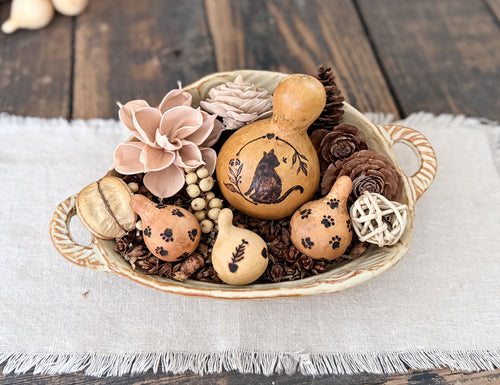 Decorative cat gourd is nestled in with smaller pet themed gourds, woods flowers, dried botanicals and twig ball in a ceramic bowl with handles. 