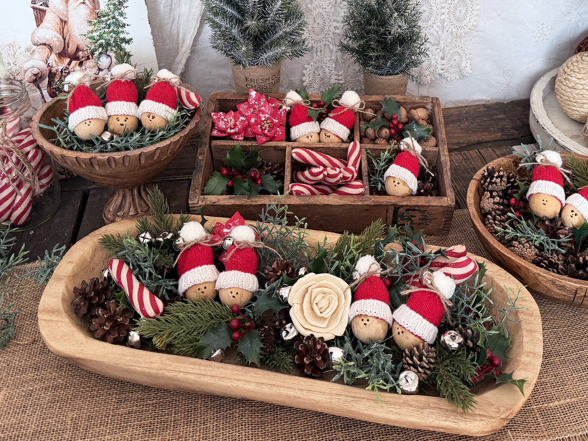 A collection of farmhouse bowl filler decorating ideas. Included a round teak bowl, a long wooden dough bow, and a vintage wooden box. All hold an assortment of greenery, pine cones and mini Santa gourd figures. 