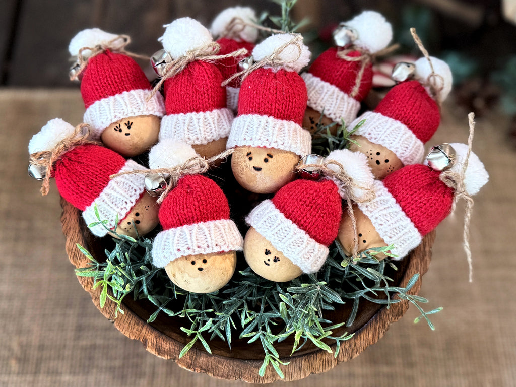 Several mini gourds with red Santa Claus hats on, arranged in a round wood bowl.  The hats have white pom-poms and silver jingle bells. Faces vary and are wood burned onto the gourd shell. 