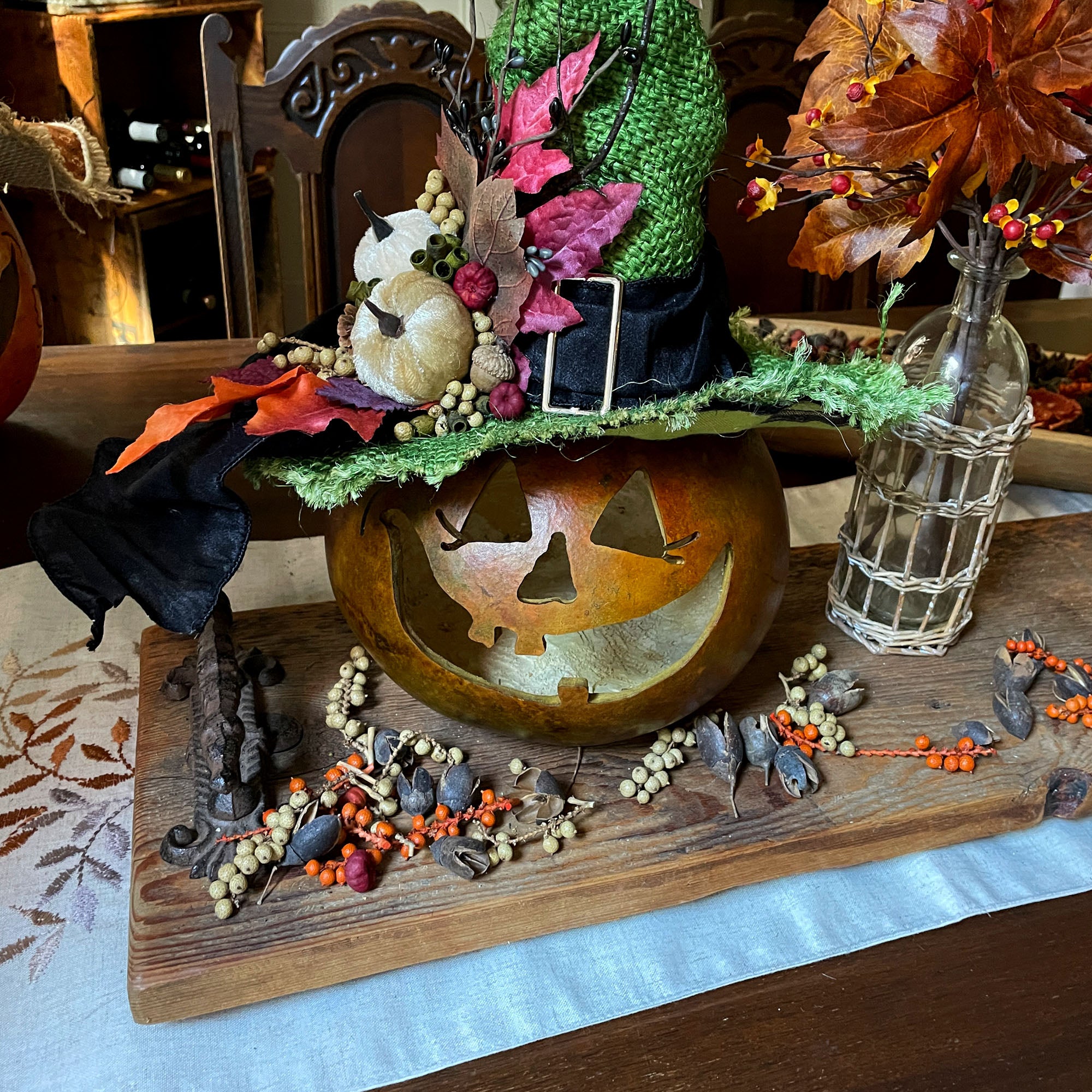 A Halloween table centerpiece with a wood riser, dried pod table scatter and a carved pumpkin witch with a green burlap hat.