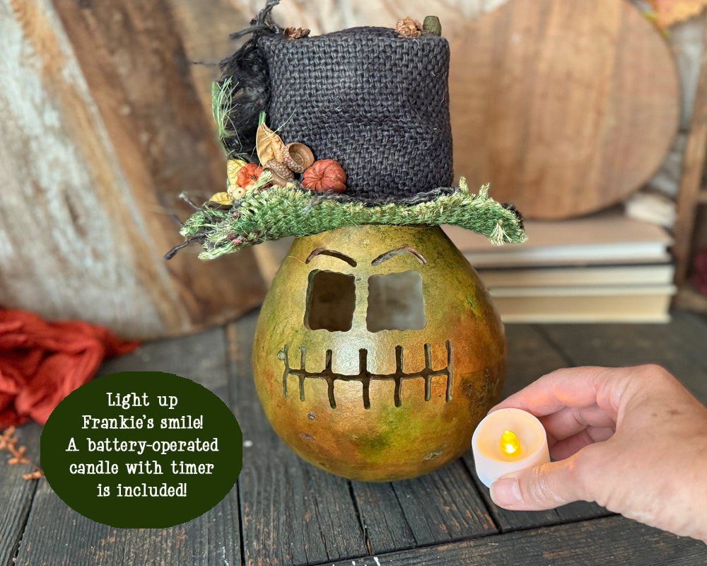 Pumpkin gourd named Frankie. Gourd shell is green with carved face. Top hat made from burlap. Hand holding batter-operated candle