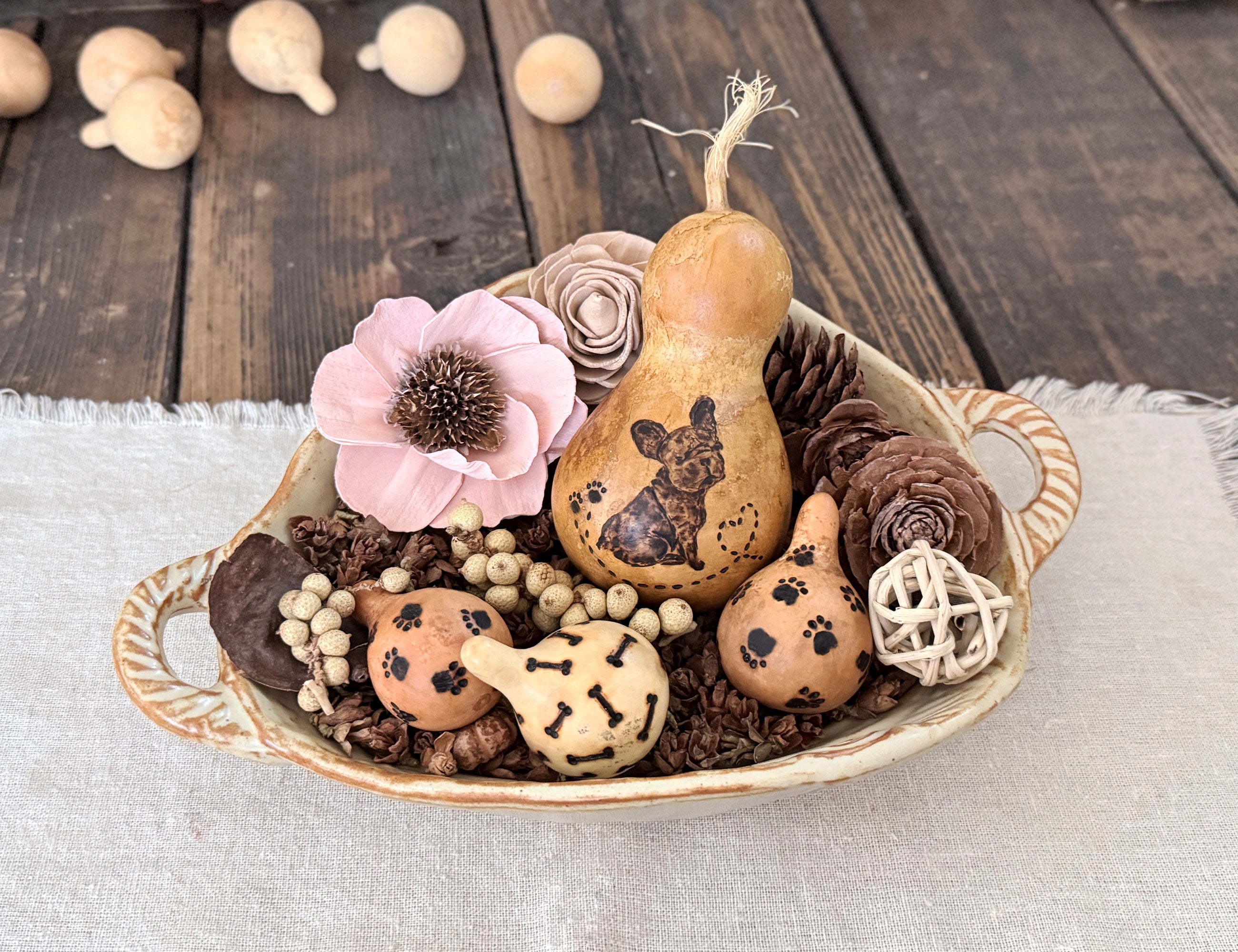Decorative arrangement with dog gourds, flowers, and pinecones in a bowl on a wooden surface.