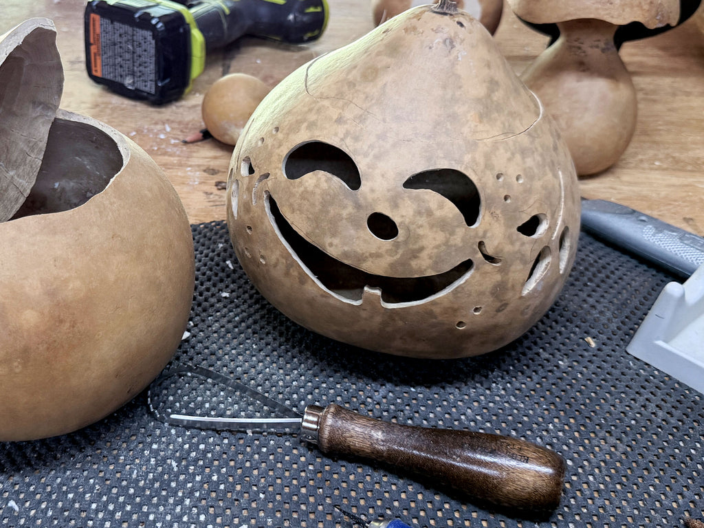 A carved natural gourd pumpkin with a smiling jack-o'-lantern face, displayed on a workbench alongside carving tools and other gourd projects.