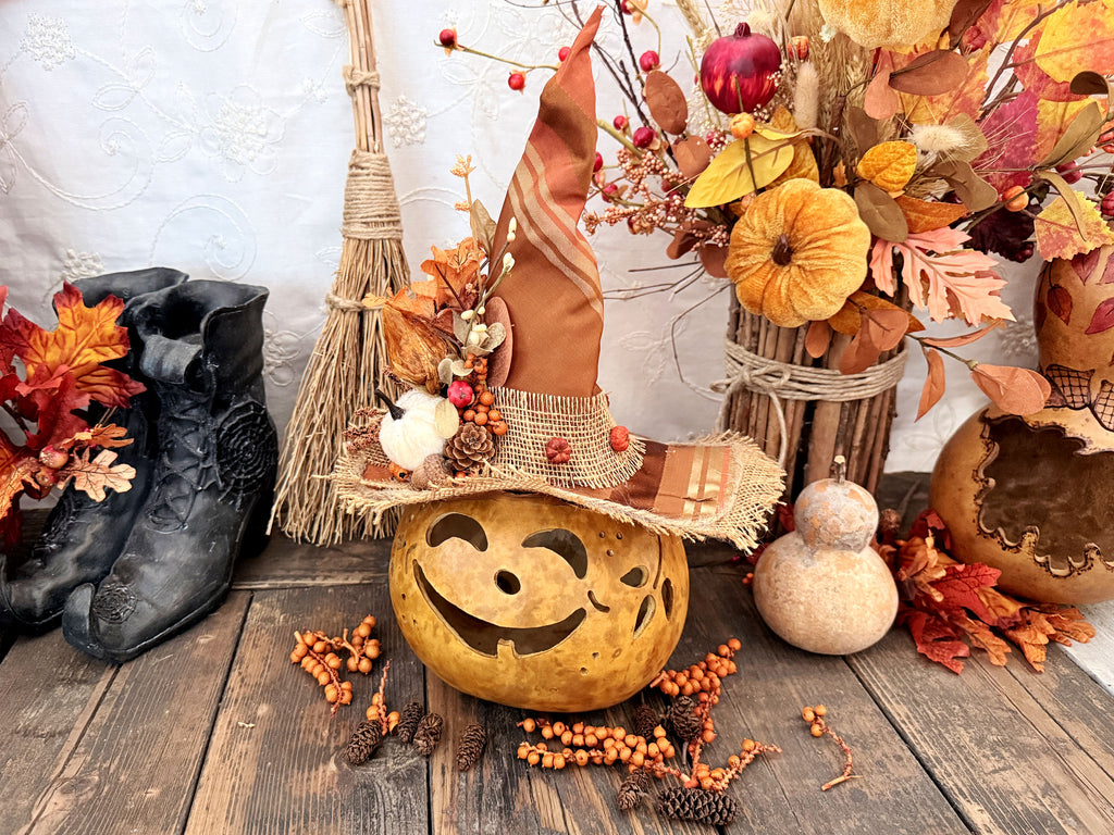 An autumn-themed decorative display featuring a carved pumpkin with a witch hat adorned with orange fabric, pinecones, and faux berries, surrounded by fall leaves, a broom, black boots, and a gourd on a rustic wooden surface.