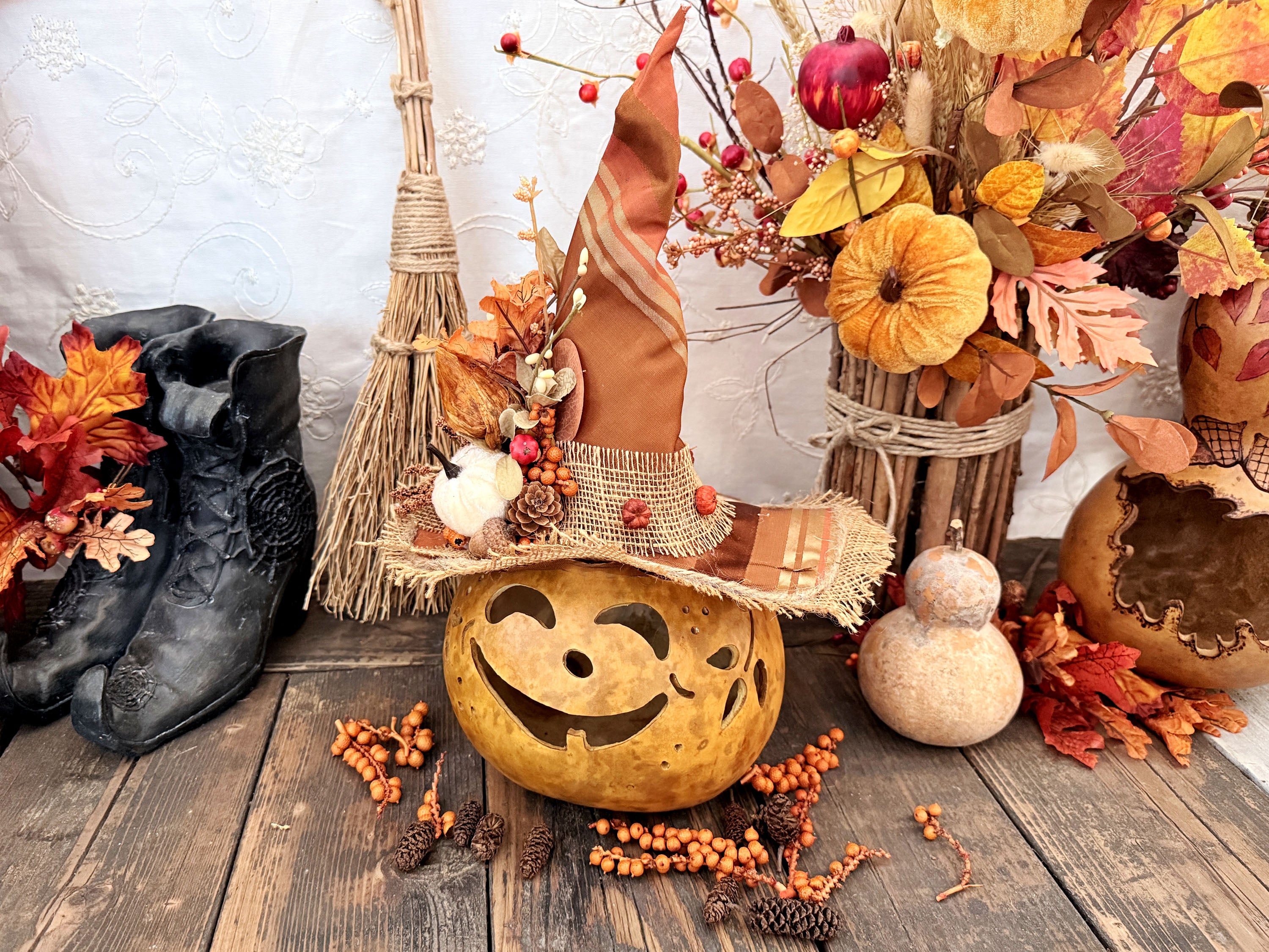 An autumn-themed decorative display featuring a carved pumpkin with a witch hat adorned with orange fabric, pinecones, and faux berries, surrounded by fall leaves, a broom, black boots, and a gourd on a rustic wooden surface.