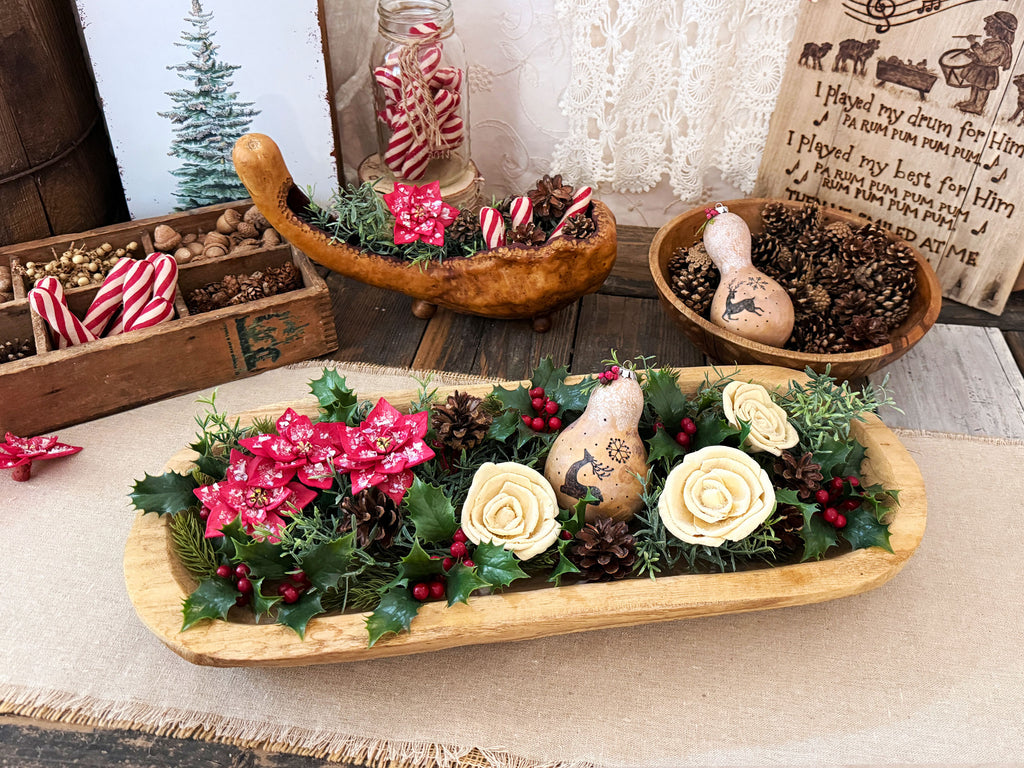A wooden dough bowl holds a holiday arrangement of greenery, and snowy winter flowers. Other arrangements are in the back with a Little Drummer Artwork sign.