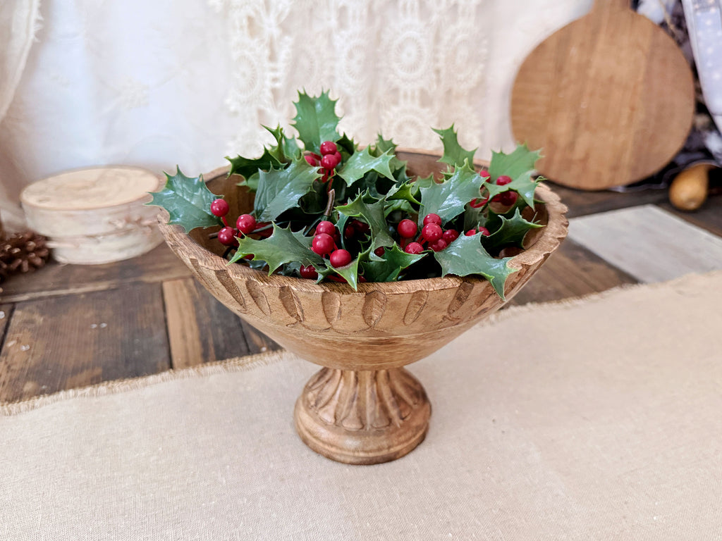 A round wood bowl holds an arrangement of realistic holly berry greens. 