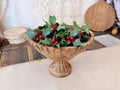 A round wood bowl holds an arrangement of realistic holly berry greens. 