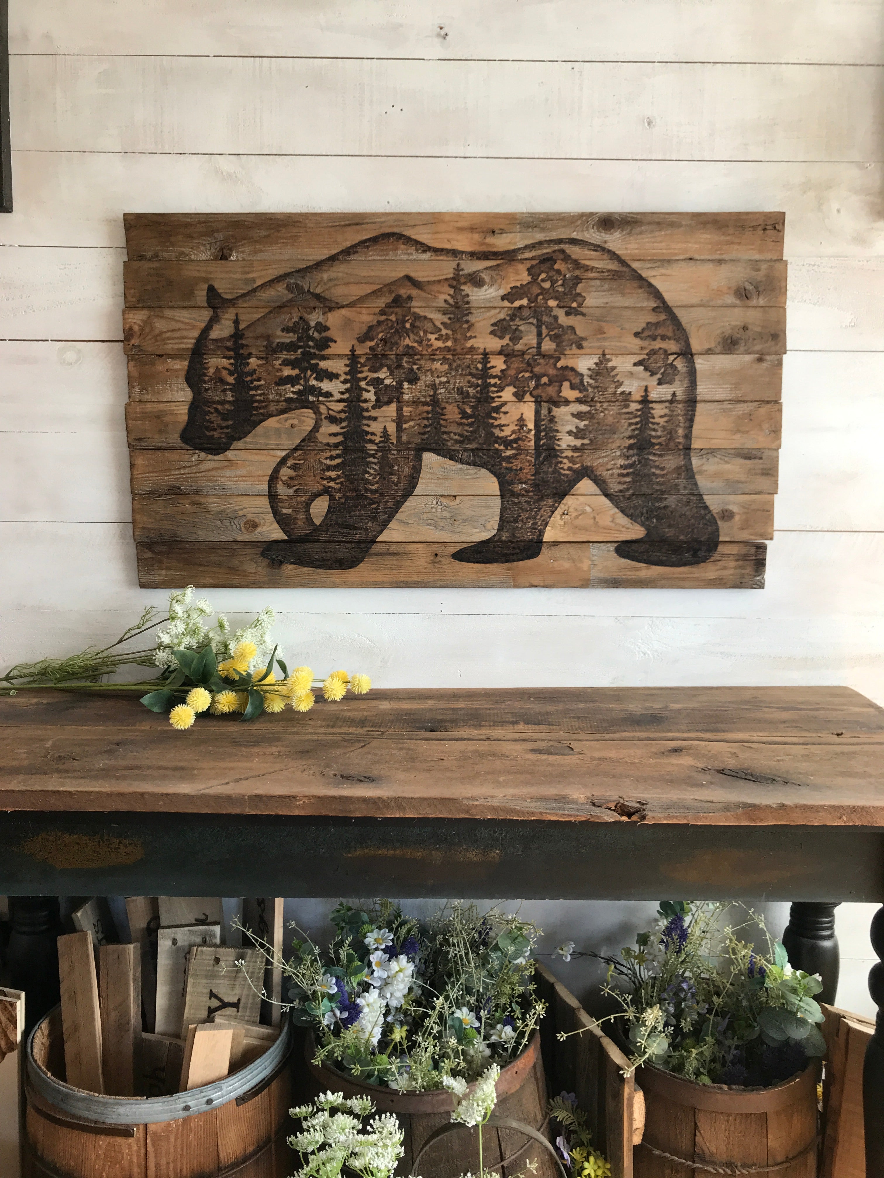 Wooden wall art of a bear with trees on a light wooden panel, next to a decorative setup with flowers and a barrel.