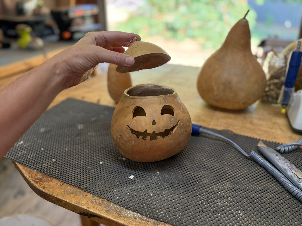Person holding a small gourd with carved face on a table with other gourds.