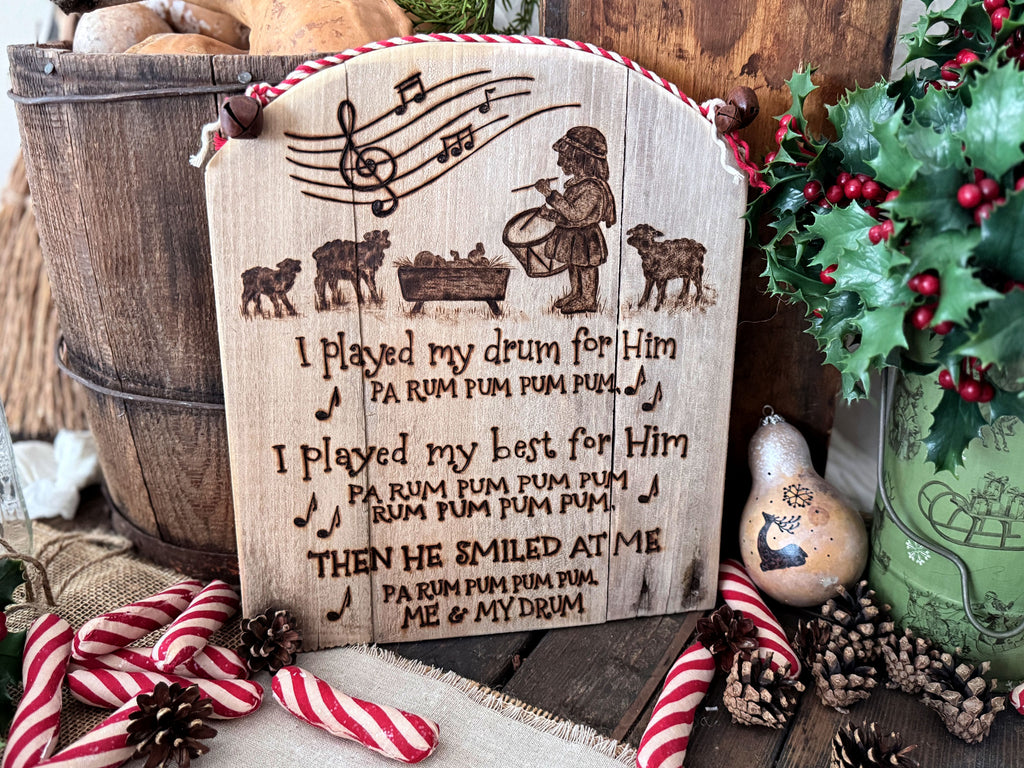 A wood wall sign made from upcycled wood with a rounded top. Artwork features the Little Drummer Boy standing near the baby Jesus with sheep near. Musical notes are at the top, with words from the song lyrics below. The artwork has  been wood burned onto the wood.  The top has red and white cord attached with rusty jingle bells. 