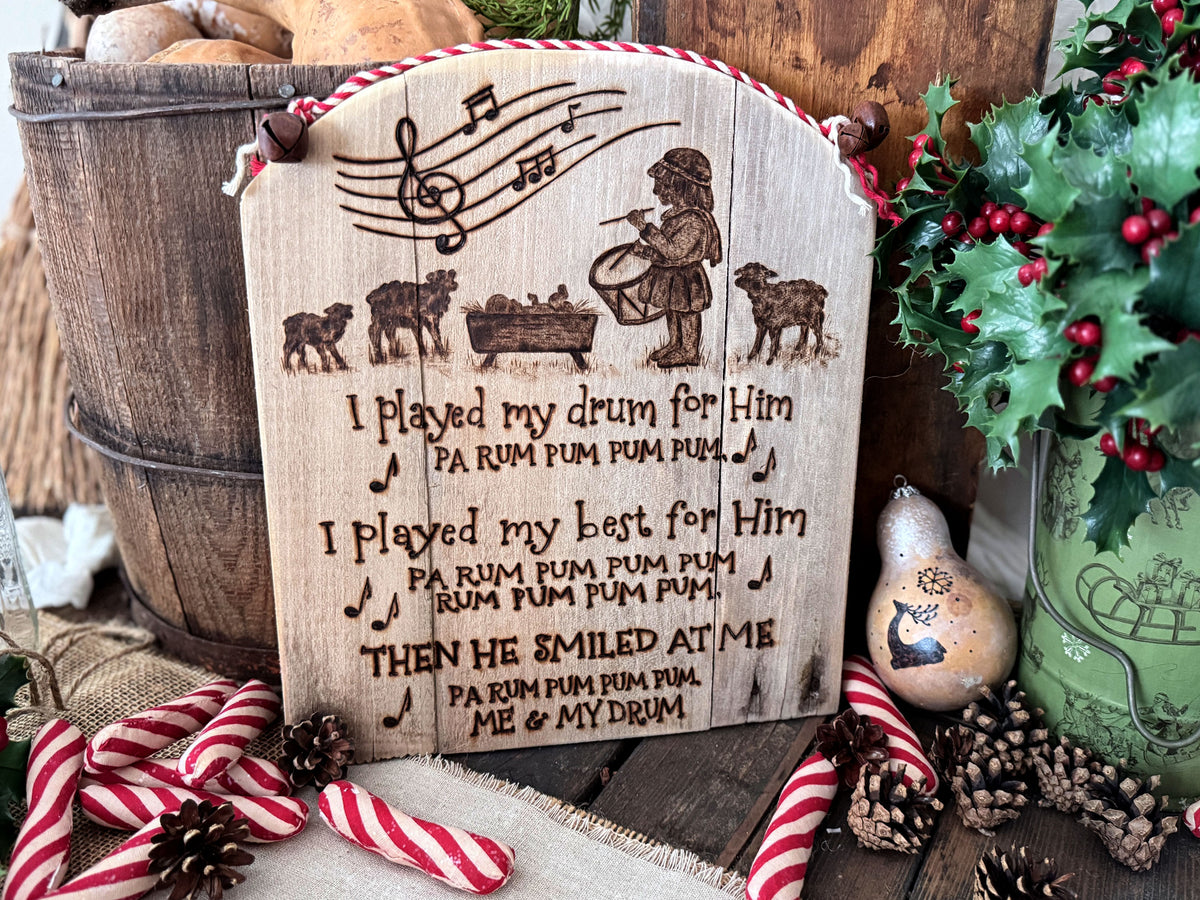 A wood wall sign made from upcycled wood with a rounded top. Artwork features the Little Drummer Boy standing near the baby Jesus with sheep near. Musical notes are at the top, with words from the song lyrics below. The artwork has  been wood burned onto the wood.  The top has red and white cord attached with rusty jingle bells. 