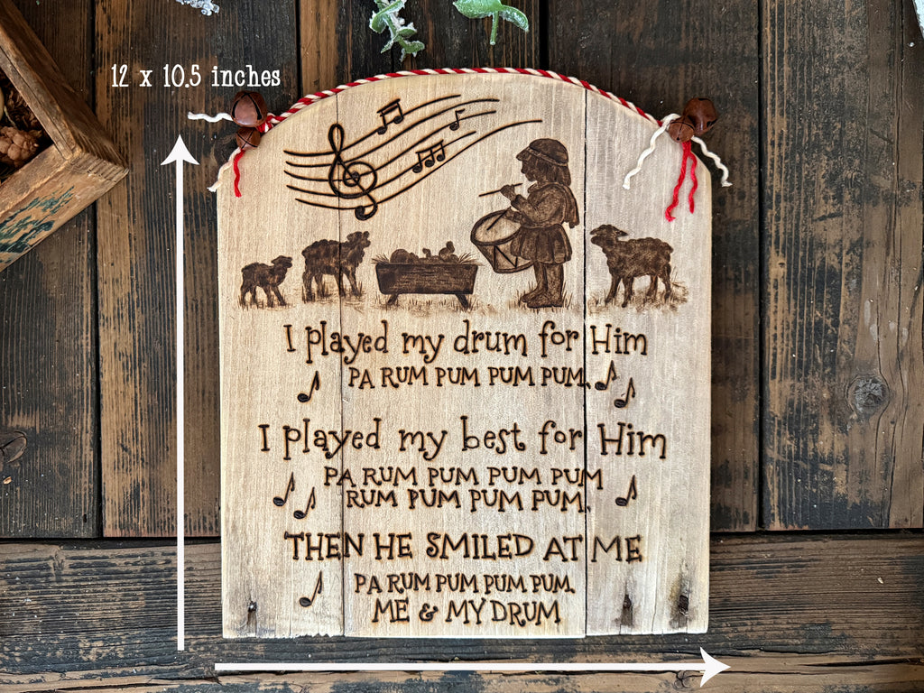 A wood wall sign made from upcycled wood with a rounded top. Artwork features the Little Drummer Boy standing near the baby Jesus with sheep near. Musical notes are at the top, with words from the song lyrics below.
Measurements with arrows show this sign is 12 by 10.5 inches. 