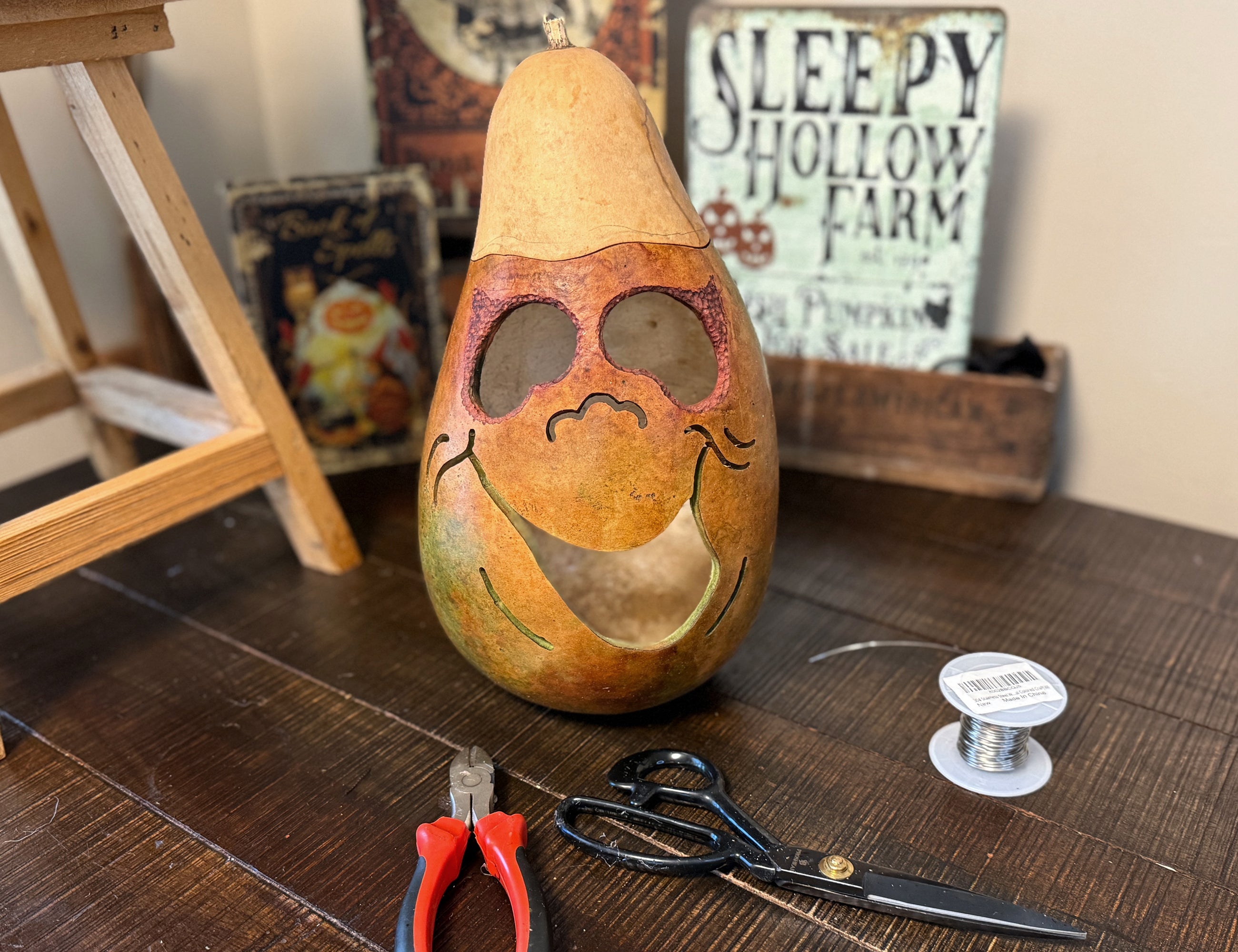 Decorative gourd with a face on a wooden surface with tools and a 'Sleepy Hollow Farm' sign in the background.