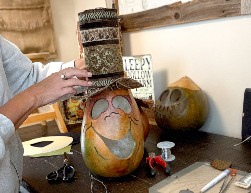 Person working on a gourd with decorative elements on a table.