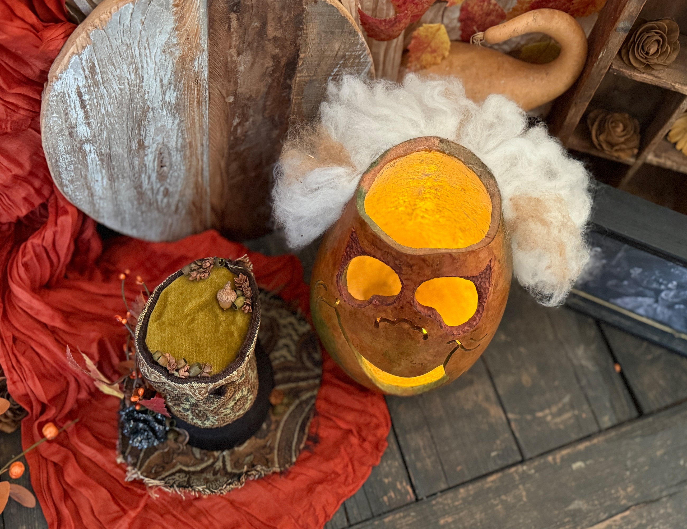 Halloween pumpkin with hat next to it. A gourd with a face, and a red fabric on a wooden surface.