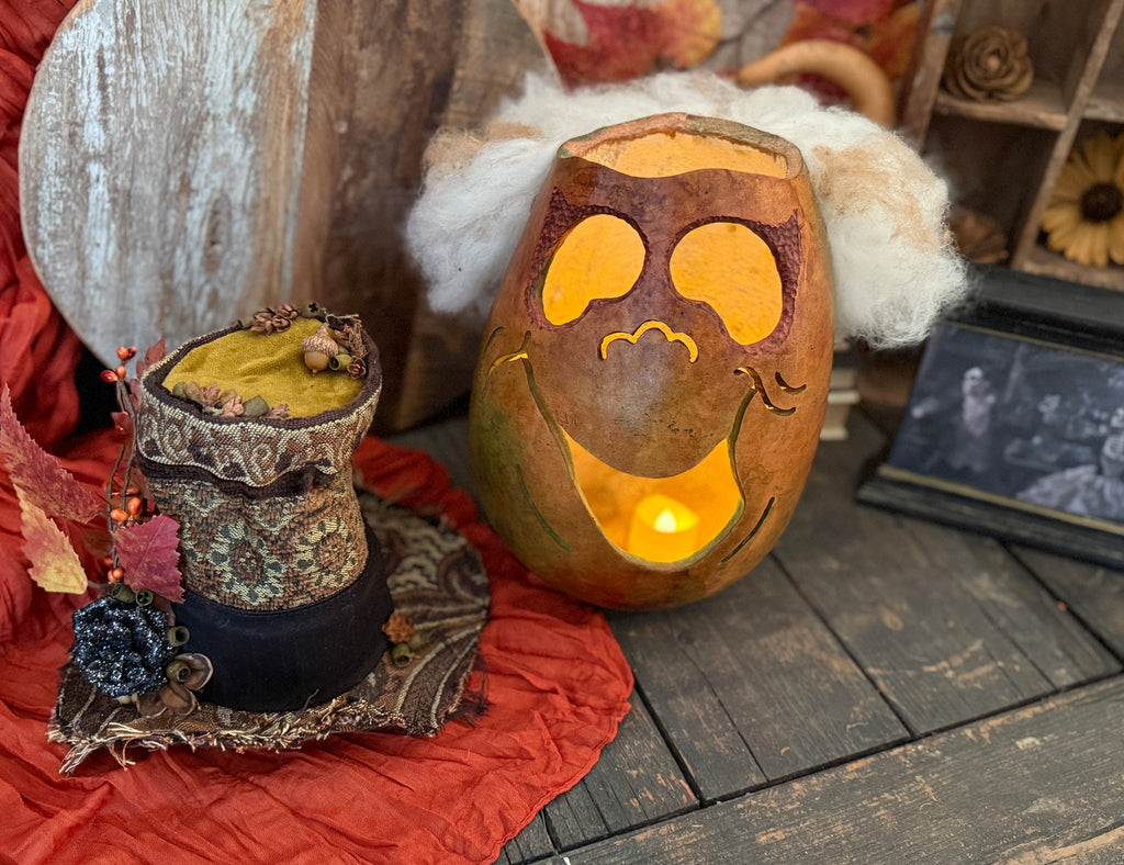 Decorative pumpkin with whimsical face carving on a rustic wooden surface with pumpkins and foliage. Top hat sits near.