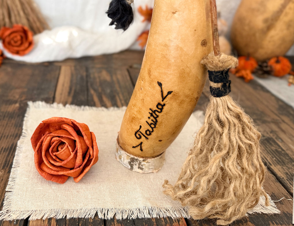 A decorative carved gourd with the name 'Tasitha' written on it, styled as a broom with a jute tassel, alongside an orange fabric rose on a rustic wooden surface.