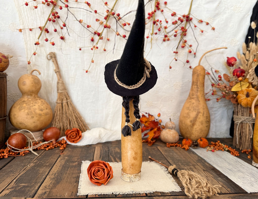 A decorative Halloween display featuring a black witch with a pointy hat with braided black hair hanging down, set on a rustic wooden table surrounded by autumn props including gourd sculptures, dried leaves, and berries.