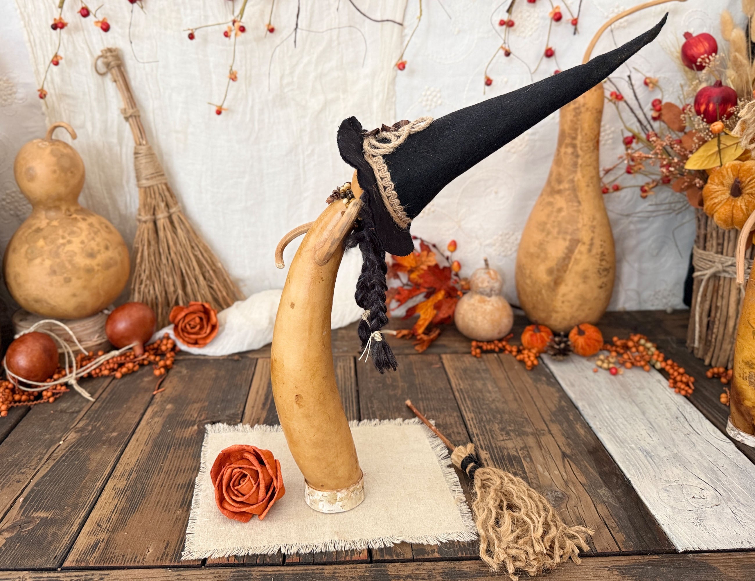 A primitive witch gourd leaning back with a black hat and braids hanging down. Surrounded by autumn props including pumpkins, a small broom, orange roses, and berries on a rustic wooden surface.