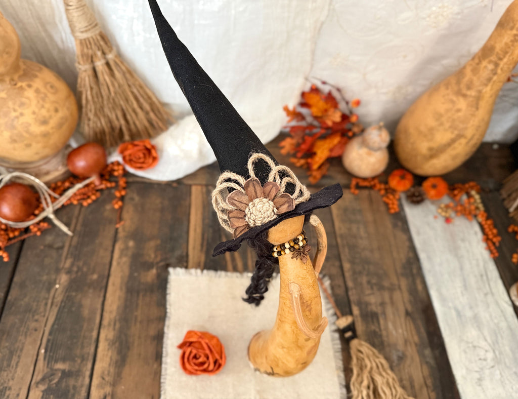 A decorative Halloween figurine shaped like a witch with a tall black hat adorned with a beige flower and black braided trim, displayed on a rustic wooden surface with autumn props including gourds, dried leaves, and a broom.