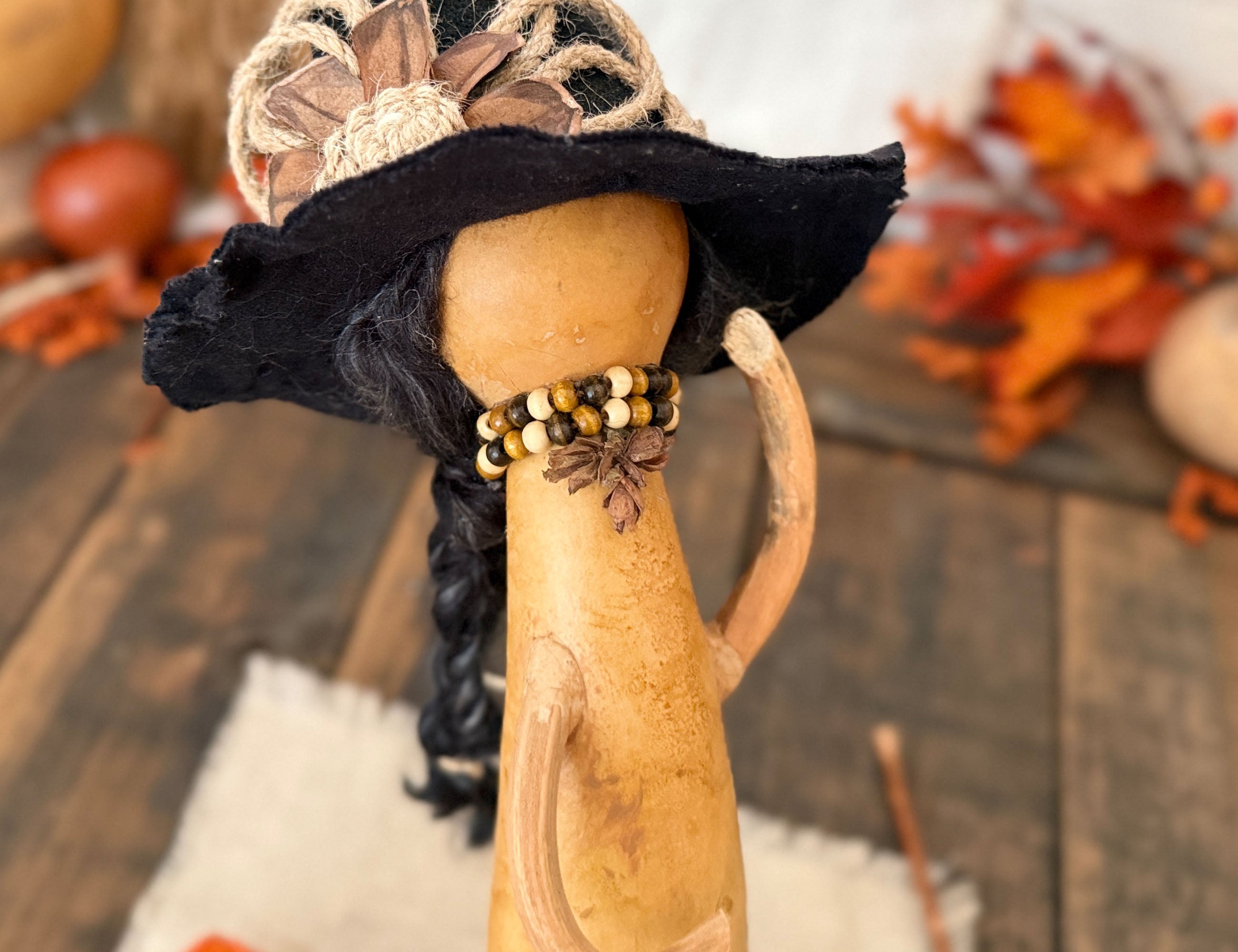 A close-up of a decorative autumn-themed display featuring a carved gourd with a black hat, braided black hair, and a beaded necklace with a star detail, set against a rustic wooden background with fall leaves.