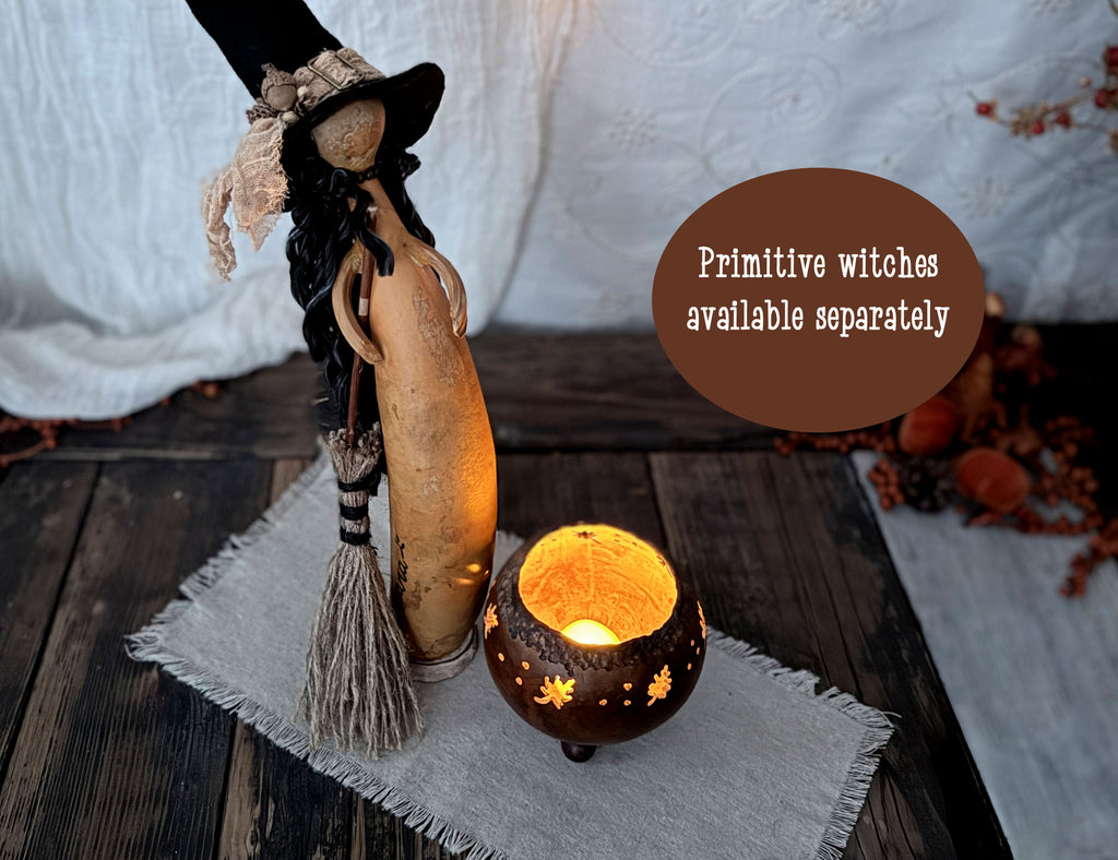 A styled tabletop scene with a carved gourd witch figure wearing a hat and holding a broom, next to a glowing carved pumpkin candle holder. A brown circular text overlay reads 'Primitive witches available separately.