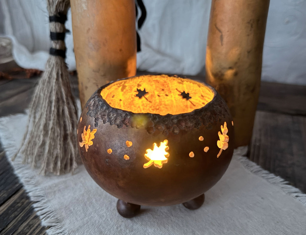 A glowing brown cauldron-shaped candle holder with cutout autumn motifs, lit with warm yellow-orange light, set on a white fringed cloth with rustic gourd witches in the background.