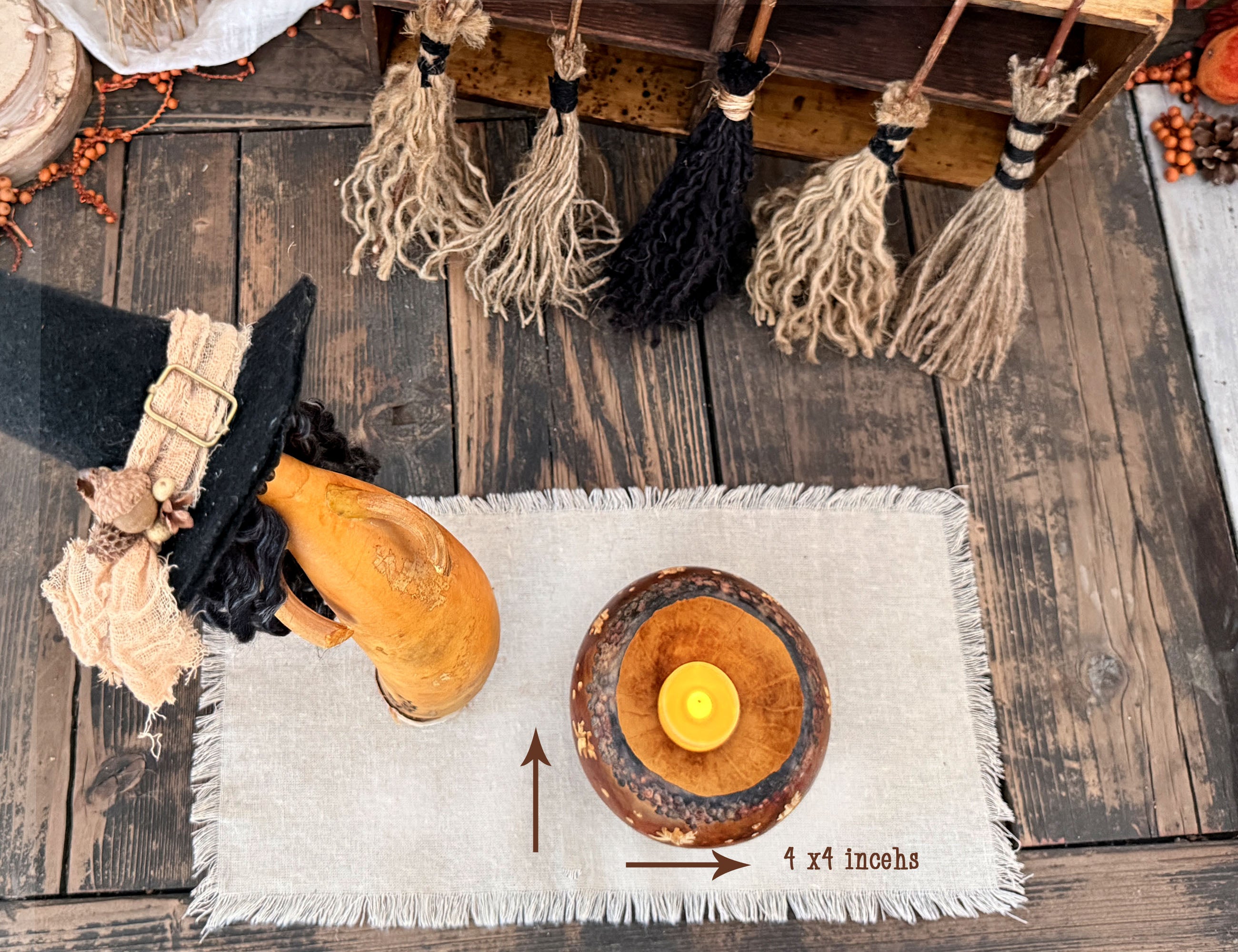 A styled flat-lay of a round gourd cauldron that acts as a candle holder with a yellow tealight candle, placed on a light beige fringed runner over a rustic dark wood surface. A witch hat and orange gourd are nearby as props, with broomsticks hanging above and measurement arrows indicating the candle holder's dimensions.