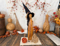 A primitive witch figurine made from dried gourds. She has a tall black pointy witchy hat and her own broom. A orange wood rose sits next her. Fall berries and gourds  sit in the background. 